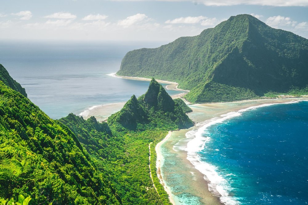 Aerial view of a lush, mountainous American Samoa coastline with turquoise reef waters and white sand beach.